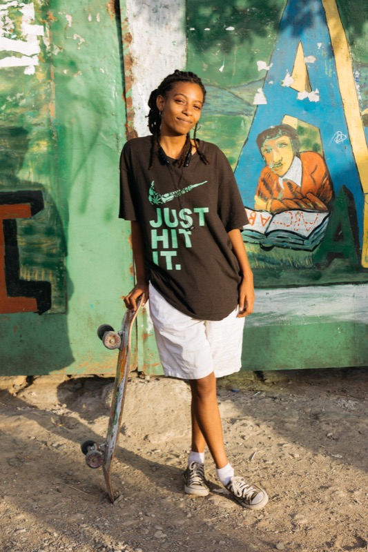 A young woman stands outdoors on a dirt surface, holding a skateboard with her right hand. She is wearing a black T-shirt with the phrase 'JUST HIT IT.' in mint green, white shorts, and tennis shoes. She has braided hair, a smile, and is wearing earp