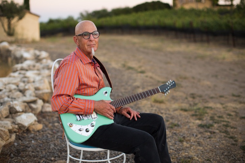 An older man with glasses, a goatee, and shaved head sitting outdoors on a white chair, holding a mint green electric guitar with a white pickguard, wearing a red striped shirt and black pants, with a rocky and natural background.