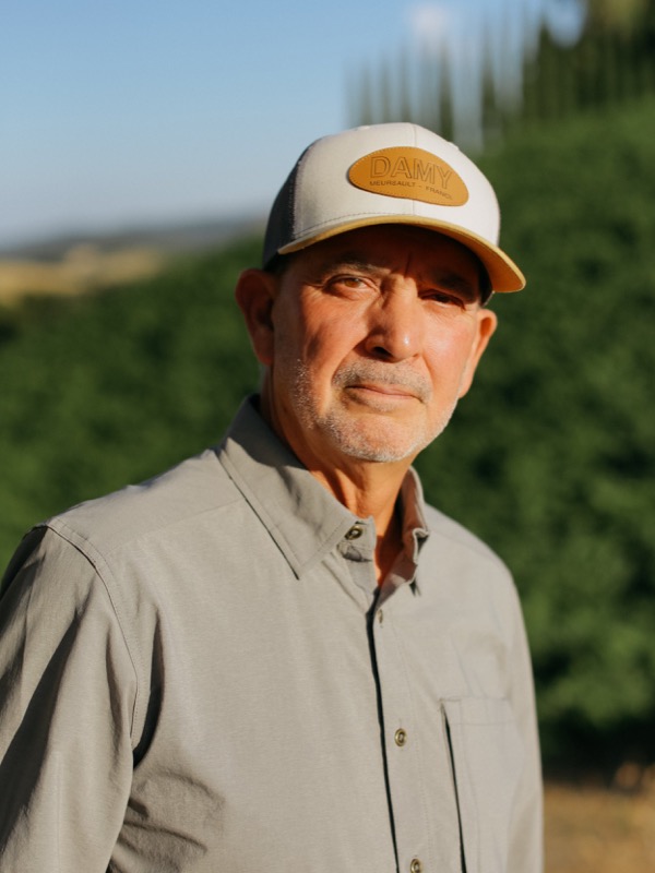 An older man with gray facial hair wearing a beige cap with a leather patch that reads "DAMY" and a light gray button-up shirt, standing outdoors with a blurred green landscape in the background.