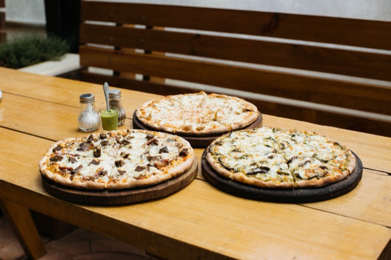 Three pizzas on wooden plates with cheese, vegetables, and meat toppings on a wooden table, with salt and pepper shakers and a small green sauce in a glass jar.