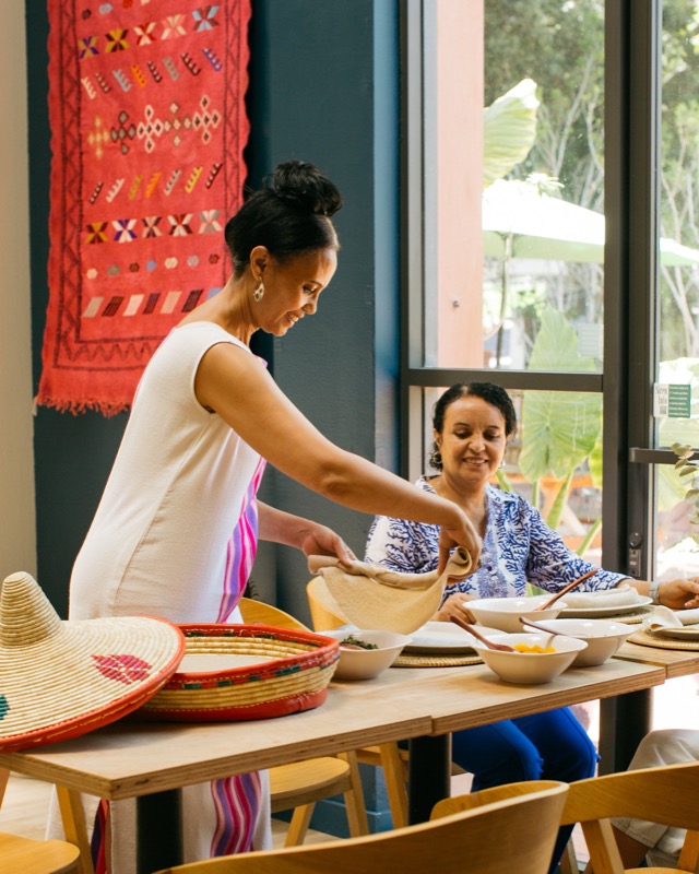 Two women preparing food or serving in a restaurant, with bowls and hand-crafted hats on the table.